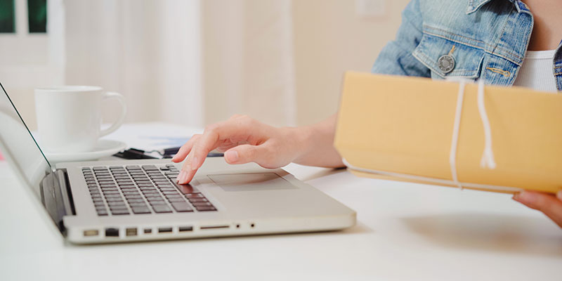 Mulher navegando no computador com caixa de encomenda em uma das mãos